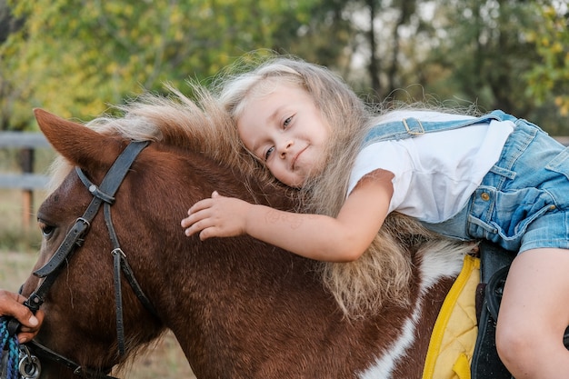 Una ragazza bionda carina è seduta su un pony in autunno.