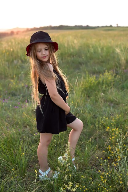 una ragazza bionda con un abito nero e un cappello panama nero si trova sotto i raggi del sole al tramonto