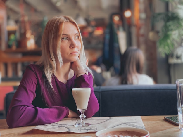 Una ragazza bionda con un maglione lilla si siede in un accogliente ristorante su un divano ed è rilassata e sorridente e beve un caffè sul suo tavolo.