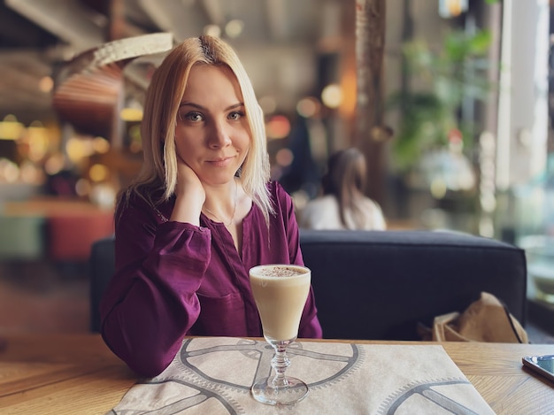 Una ragazza bionda con un maglione lilla si siede in un accogliente ristorante su un divano ed è rilassata e sorridente e beve un caffè sul suo tavolo.