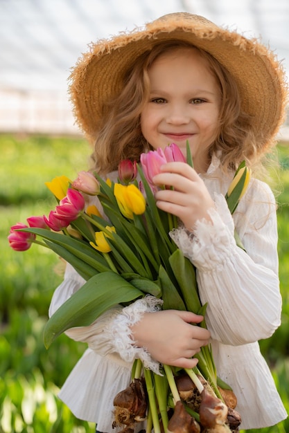 una ragazza bionda con una camicetta bianca e un cappello di paglia tiene in mano un mazzo di tulipani colorati