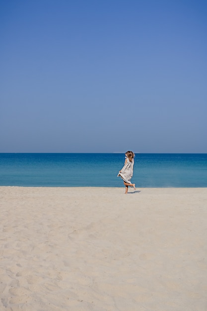 Una ragazza bionda sorridente in un maxi abito di lino con svolazzanti capelli che salta e balla su una spiaggia di sabbia