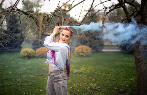 Una ragazza brillante con il trucco glitterato e le trecce africane in una camicia bluastra si rifugia nel fumo artificiale in un parco pieno di alberi verdi