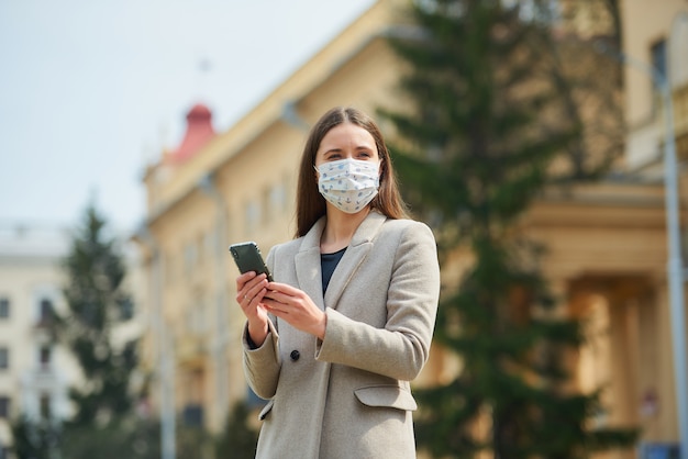 Una ragazza bruna con i capelli lunghi in una maschera medica per evitare la diffusione del coronavirus cerca notizie su uno smartphone nella vecchia strada. Una donna in maschera contro COVID-19 mantiene una distanza sociale.