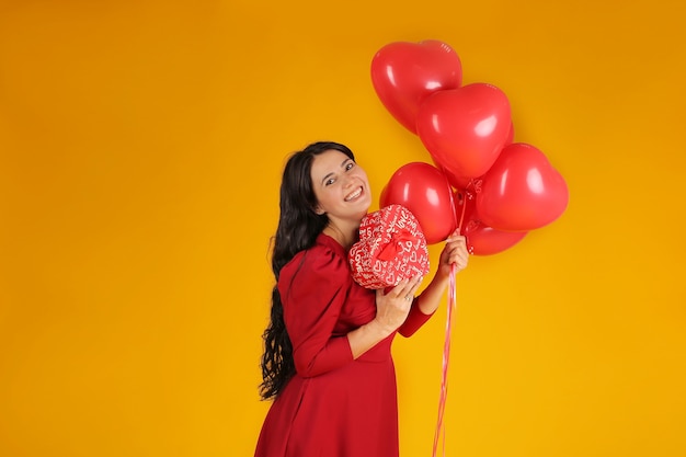 una ragazza bruna con un vestito rosso tiene in mano dei palloncini una confezione regalo a forma di cuore