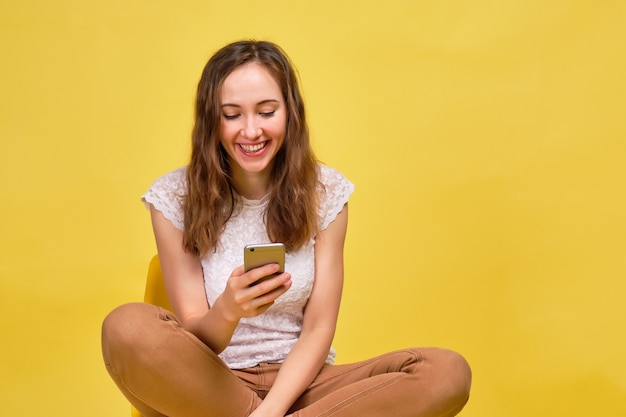 Una ragazza bruna in abiti estivi è sorridente e seduto guardando in uno smartphone su uno sfondo giallo. Il concetto di invio di sconti sugli acquisti.