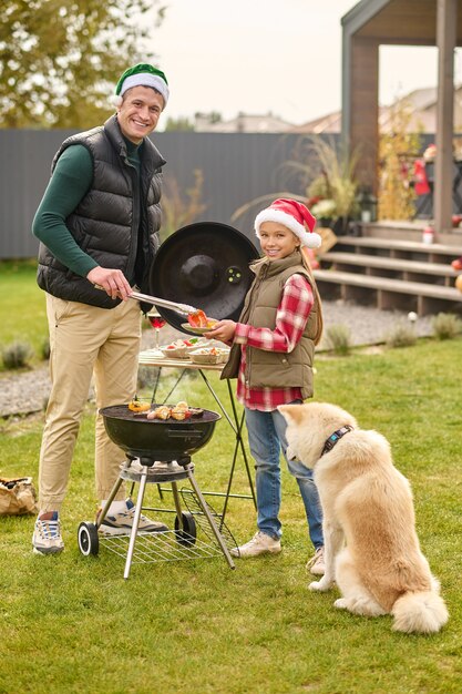 Una ragazza carina con un cappello da Babbo Natale in piedi sulla griglia all'aperto mentre suo padre cucina