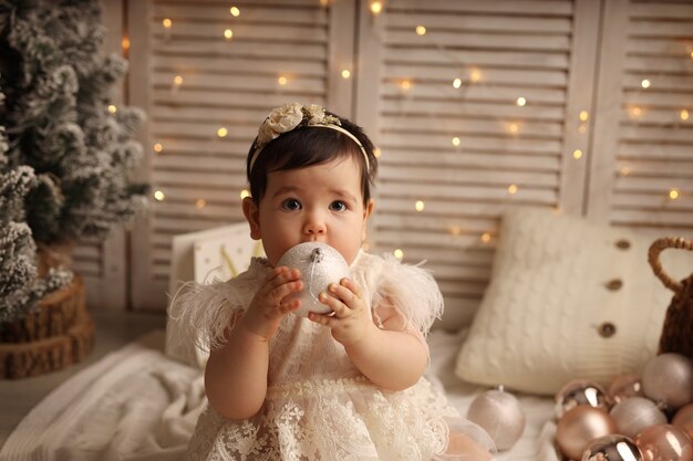 Una ragazza carina dalla pelle scura è seduta su una coperta a maglia bianca e gioca con i giocattoli dell'albero di Natale