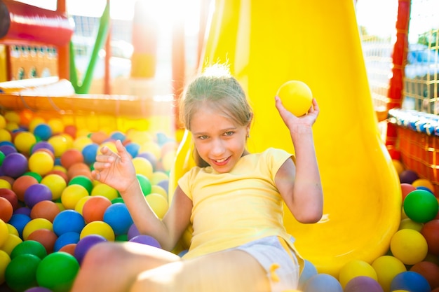 Una ragazza carina e divertente si siede in un parco giochi con attrezzature morbide e luminose e lancia palline colorate verso la telecamera