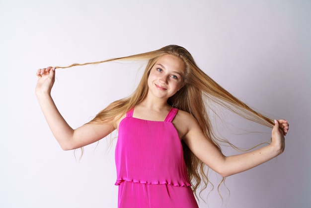 Una ragazza carina e felice di aspetto caucasico in abiti rosa su un muro bianco gioca con i suoi lunghi capelli