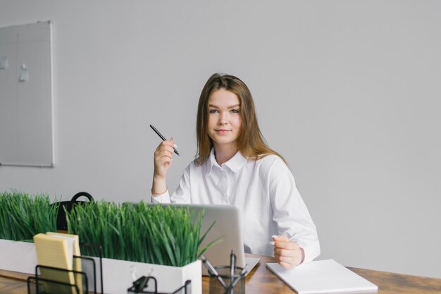 Una ragazza carina è seduta a una scrivania in ufficio, lavora con un laptop e un blocco note e guarda
