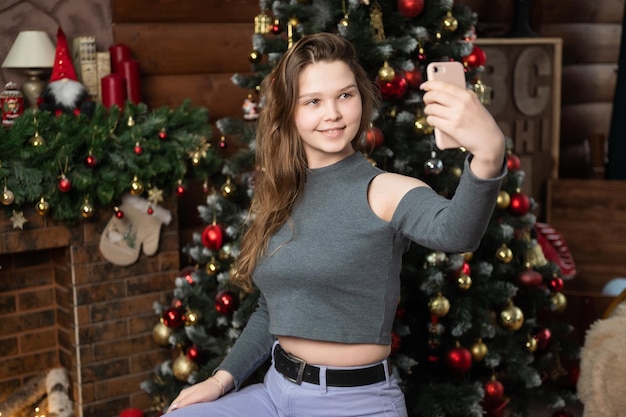 Una ragazza carina è seduta vicino all'albero di Natale con un telefono e si fa un selfie