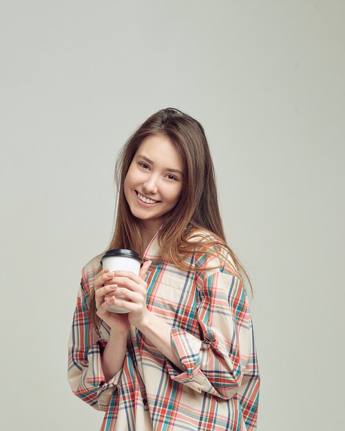 Una ragazza carina, in abiti casual, sorride dolcemente e tiene un bicchiere di caffè in una confezione usa e getta.