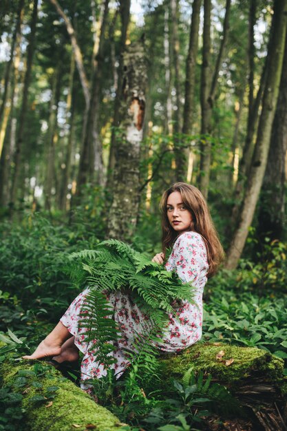 Una ragazza carina in un abito floreale è seduta con un bouquet di felci nella foresta.