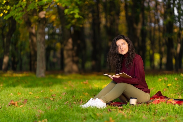 Una ragazza carina legge un libro e beve caffè su un prato verde in un parco autunnale. Atmosfera autunnale. Un luogo accogliente per stare da solo con te stesso.