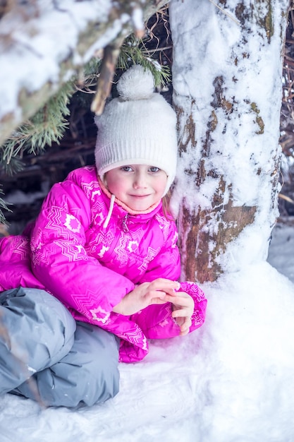 Una ragazza caucasica di 5 anni scalda le mani in una capanna da rami di conifere nella foresta invernale, giocando all'aperto in inverno