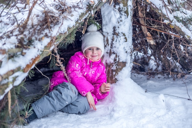 Una ragazza caucasica di 5 anni si nasconde in una capanna da rami di conifere nella foresta invernale, giocando all'aperto in inverno