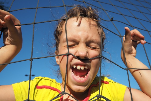 Una ragazza caucasica di 8 anni sta dietro una rete sportiva e urla Sport di pallavolo che divide la rete contro il cielo blu primo piano Sport all'aperto