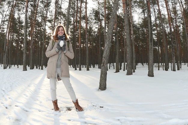 Una ragazza caucasica giovane e allegra in un cappotto marrone tiene una palla di neve in una foresta innevata in inverno. Foto Fisheye