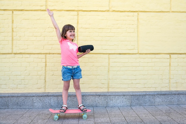 Una ragazza cavalca uno skateboard con un altoparlante di musica bluetooth contro un muro di mattoni gialli