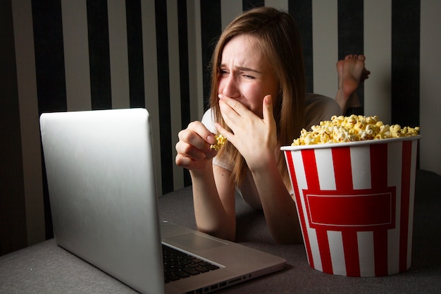 Una ragazza che guarda un programma televisivo di notte su un laptop e mangia popcorn, usa un computer mentre giace sul divano