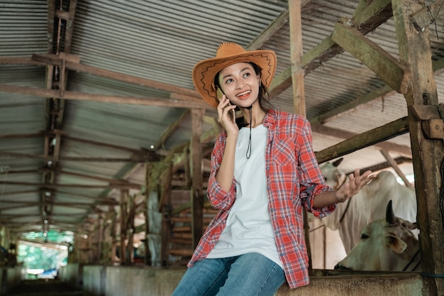 Una ragazza che indossa un cappello effettua una chiamata utilizzando uno smartphone sullo sfondo di una staccionata in legno di fattoria di mucche