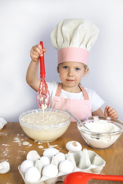 Una ragazza con cappello e grembiule da chef prepara l'impasto, frusta tra le mani, uova, farina sul tavolo, bianco