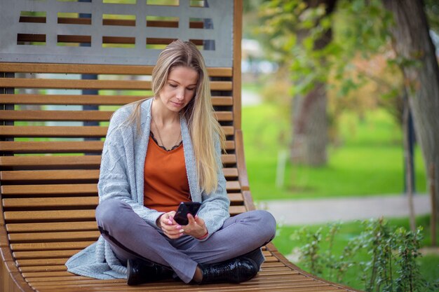 Una ragazza con i capelli lunghi e in una giacca grigia si siede su una panchina nel parco e gioca su uno smartphone