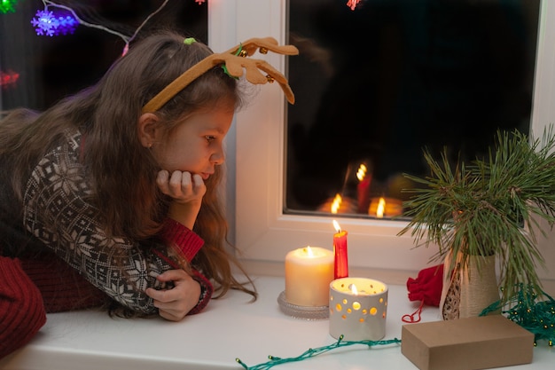 Una ragazza con le corna di cervo si siede sul davanzale della finestra a Natale e guarda le candele