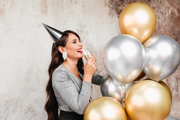 Una ragazza con palloncini sta ridendo sullo sfondo di una parete decorativa Una bella donna felice per il compleanno di una festa di Capodanno divertendosi con in mano un bicchiere di champagne