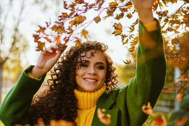 Una ragazza con piccoli capelli ricci con un bel sorriso dai denti bianchi tocca con le mani i rami di