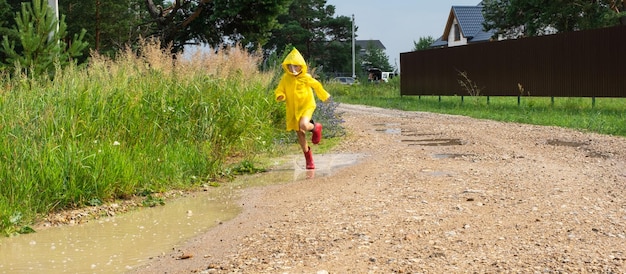 Una ragazza con stivali di gomma rossi e un impermeabile giallo corre attraverso le pozzanghere dopo una pioggia nel villaggio Estate libertà infanzia