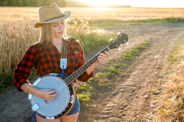 Una ragazza con un banjo sta in un campo al tramonto e canta una canzone. Stile country. Messa a fuoco selettiva.