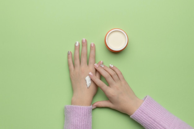 Una ragazza con un bel trucco si sta applicando la crema sulle mani da un barattolo. Disposizione piatta.