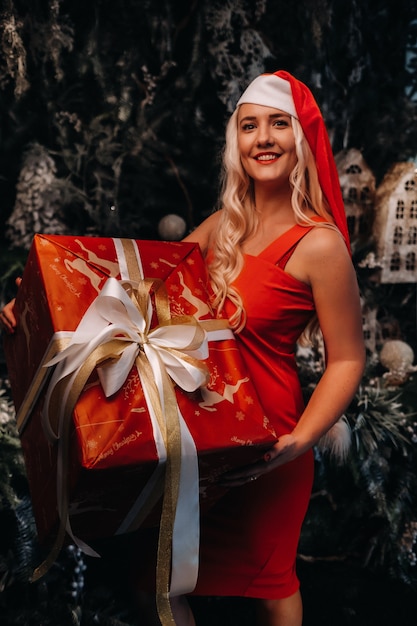 Una ragazza con un cappello da Babbo Natale con un grande regalo di Natale nelle sue mani su uno sfondo di Natale favoloso. Una donna sorridente in un vestito rosso sullo sfondo di alberi di Natale e piccole case