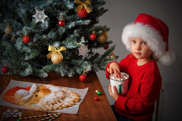 Una ragazza con un cappello da Babbo Natale con una tazza accanto a una torta di Natale a forma di Babbo Natale e decorazioni