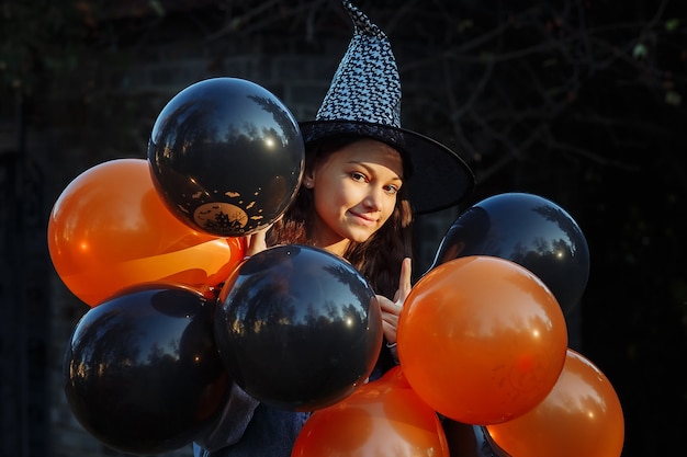 una ragazza con un cappello da strega con palloncini neri e arancioni in mano si sta preparando per Halloween