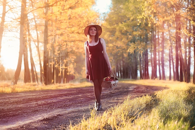 Una ragazza con un cappello durante una passeggiata nel parco. Una ragazza con un cesto cammina in autunno. La ragazza sta camminando lungo la strada al tramonto.