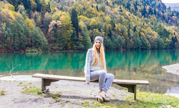 Una ragazza con un cappello è seduta su una panchina sullo sfondo di una foresta autunnale e di un lago