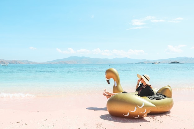 Una ragazza con un cappello estivo seduto su un cigno gonfiabile mentre si gode la vista del mare in una spiaggia di sabbia rosa