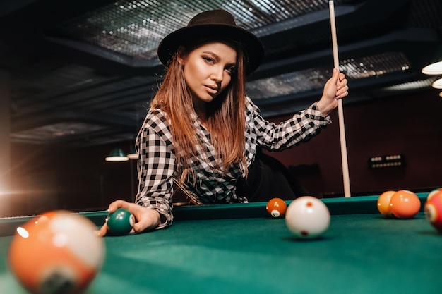 Una ragazza con un cappello in un club di biliardo con una stecca e le palle in mano. Giocare a biliardo.