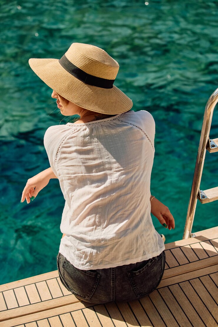 Una ragazza con un cappello si siede sul bordo di una barca a vela e gode della vista e del colore azzurro del mare