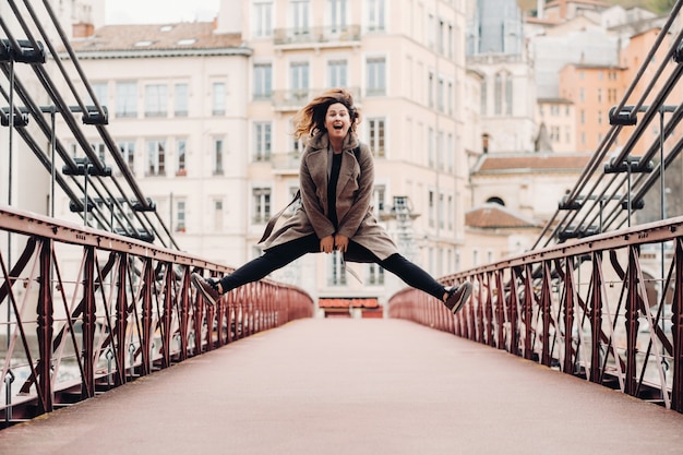 Una ragazza con un cappotto e i capelli sciolti salta emotivamente su un ponte nella città vecchia di Lione. Francia. Ragazza con un cappotto in Francia.