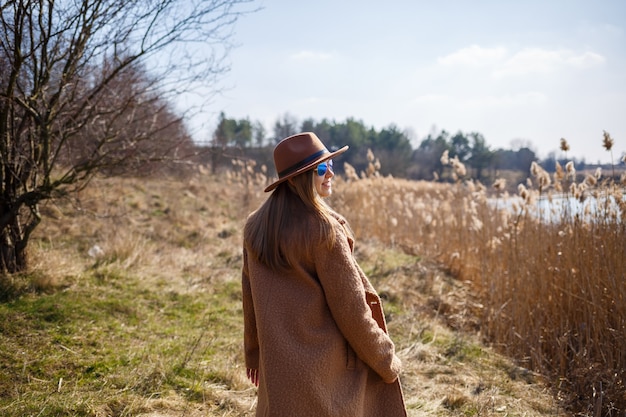 Una ragazza con un cappotto marrone, cappello e occhiali cammina in un parco con un lago sotto il sole splendente. Si rallegra della vita e sorride. L'inizio della primavera