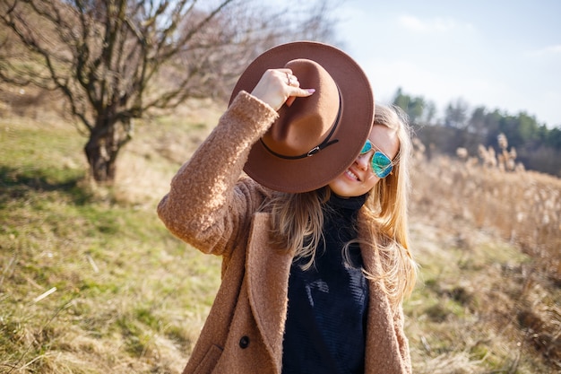 Una ragazza con un cappotto marrone, cappello e occhiali cammina in un parco con un lago sotto il sole splendente. Si rallegra della vita e sorride. L'inizio della primavera