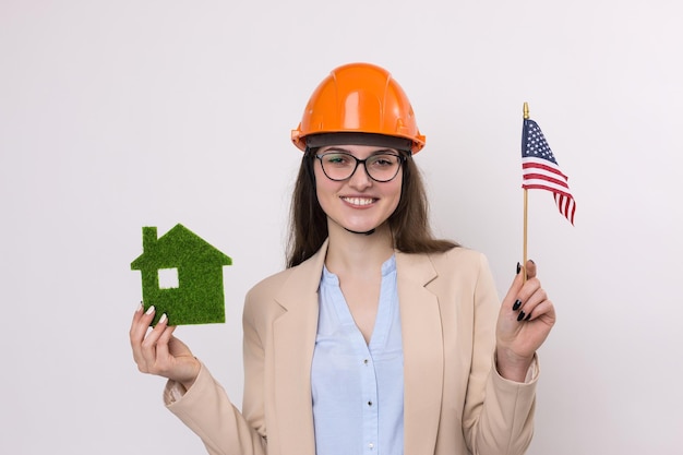 Una ragazza con un casco da costruzione e una bandiera americana tiene una casa ecologica verde