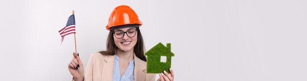 Una ragazza con un casco da costruzione e una bandiera americana tiene una casa ecologica verde