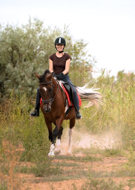 Una ragazza con un casco da equitazione cavalca un cavallo pezzato al trotto in un campo tra cespugli verdi verso il fotografo