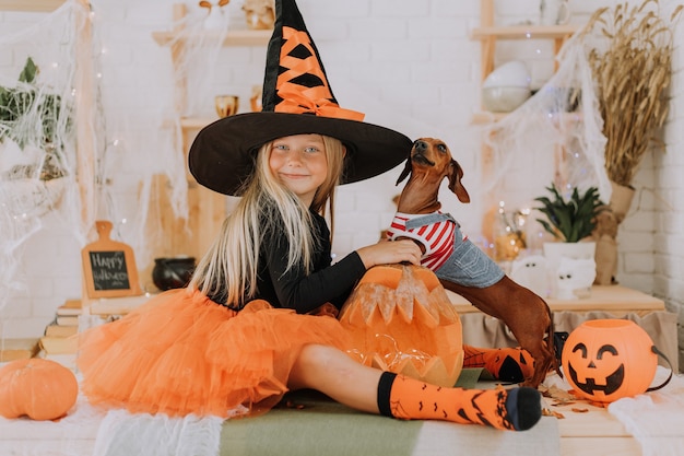 una ragazza con un costume da strega di Halloween e un piccolo bassotto con una tuta da cane sono seduti sul pavimento