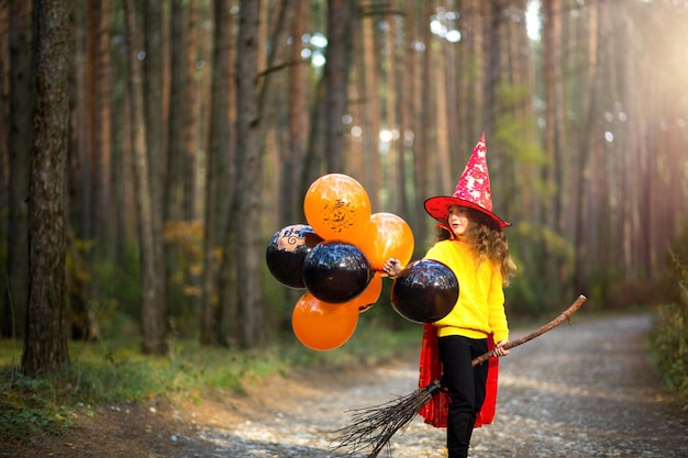 Una ragazza con un costume da strega e un cappello su una scopa con palloncini arancioni e neri sta giocando nella foresta autunnale andando a una festa di Halloween